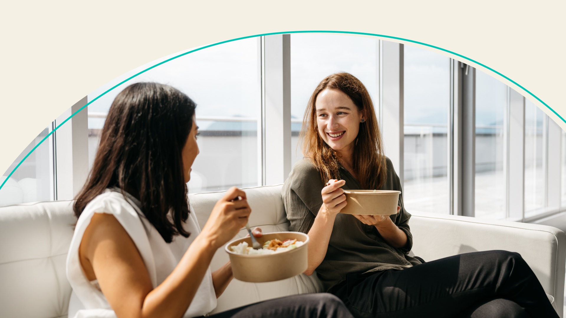 Two women sitting on a couch eating lunch together at work